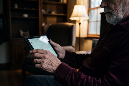 A retired elderly man with a grey beard sits comfortably in his warm living room holding a digital tablet that displays medical heart rate graphs and health statistics on the illuminated screen.の素材