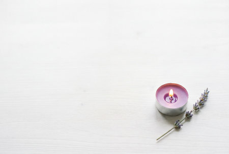 A single lit purple tealight candle sits next to a delicate sprig of blooming lavender on a white painted wooden surface with plenty of empty space for text or design overlays.の素材