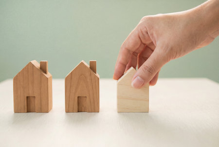 A hand carefully places a light wooden house model in a line with two darker wooden houses on a clean table to symbolize property investment and real estate decisions.の素材