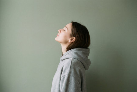 A relaxed young woman wearing a comfortable grey hooded sweatshirt tilts her head back in profile with eyes closed against a solid muted green background.の素材