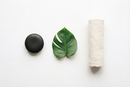 A minimalist flat lay composition featuring a smooth black massage stone beside a fresh green monstera leaf and a rolled beige linen towel arranged neatly in a horizontal row on a white background.の素材