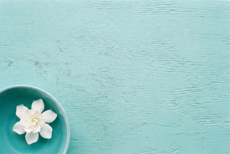 A fresh white gardenia blossom floats in water within a blue ceramic bowl placed in the corner of a weathered light turquoise wooden background with ample copy space.の素材