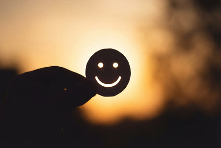 A silhouette of a hand holds a circular paper cutout with a happy smiley face against a blurred background of warm golden sunset light shining brightly through the eyes and mouthの素材