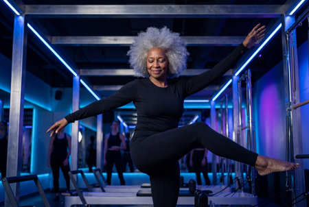 Active senior black woman with gray afro balances on one leg performing graceful pilates pose in dark studio illuminated by blue and purple neon lights.の素材