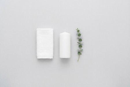 A clean minimalist arrangement featuring a neatly folded white towel next to a white pillar candle and a fresh eucalyptus branch on a plain light gray background.の素材