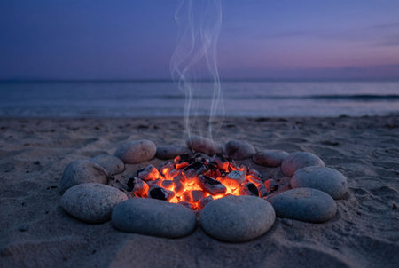 A peaceful scene showing red glowing coals and smoke rising from a campfire ring made of grey stones on a sandy beach with the calm blue ocean and purple sky visible in the background at dusk.の素材