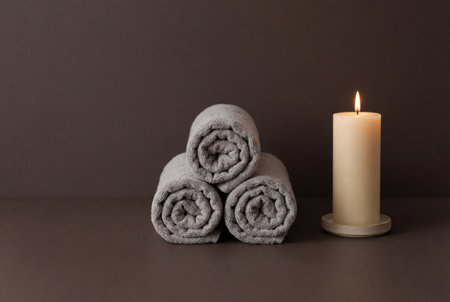 A minimalist spa setting featuring a pyramid stack of three rolled grey cotton towels placed next to a single burning beige candle on a smooth dark brown surface for a peaceful wellness atmosphere.の素材