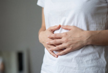 Unrecognizable woman holds her hands on her belly feeling abdominal pain or discomfort caused by menstruation or digestive issues while standing indoors.の素材