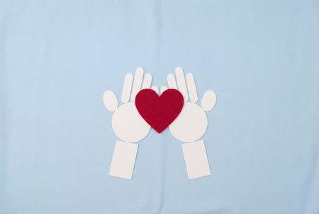 A top view flat lay composition showing stylized white paper hands gently holding a red heart shape on a light blue surface to represent concepts of love, charity, donation, healthcare and kindness.の素材