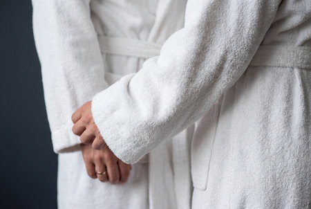A close view of a married couple wearing soft white terry cloth bathrobes standing together and embracing with a focus on their clasped hands and wedding ring.の素材