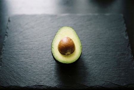 A fresh ripe avocado half with a large brown pit sits in the center of a dark grey slate board showing vibrant green flesh and a rough stone texture perfect for healthy food concepts.の素材