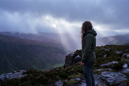 A solitary female hiker with windswept hair stands on a rocky summit overlooking a deep mountain valley where sunbeams pierce through dark storm clouds.の素材