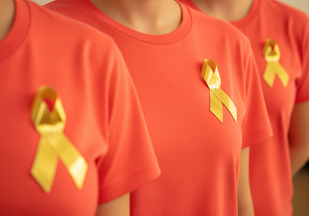 Three women standing in a line wear bright orange tshirts, each displaying a yellow ribbon pinned to the chest, symbolizing awareness, support, and solidarity for a specific charitable cause or health campaign.の素材