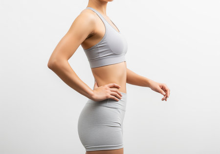 A slender woman in grey athletic sportswear is captured in a side profile studio shot, highlighting her toned abdomen, slim waist, and fit upper body against a clean white background.の素材