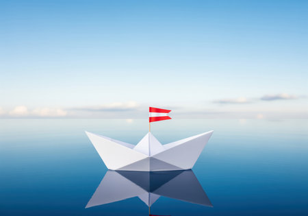 A perfectly folded white origami paper boat featuring a small red and white flag sails peacefully across the perfectly calm, reflective blue ocean surface under a bright, clear blue sky, symbolizing travel and new beginnings.の素材