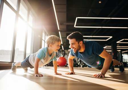 A joyful father and his young son are exercising together, performing pushups side by side on the wooden floor of a modern, sunlit fitness center, sharing a moment of bonding and healthy activity.の素材