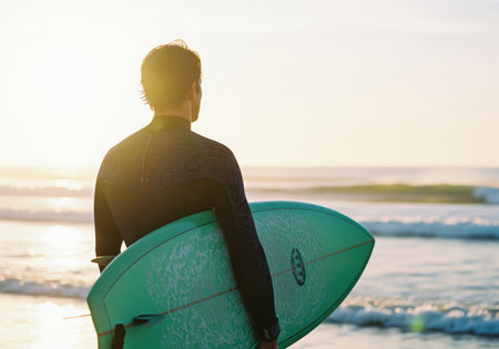 Rear view of a man in a wetsuit holding a surfboard, standing on the shore and observing the ocean waves at sunrise or sunset. ideal for themes of adventure, sport, travel, and freedom.の素材