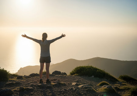 Rear view of a triumphant woman standing on a rocky mountain peak with arms wide open, silhouetted against a brilliant golden sunset over the vast ocean and distant hills. this captures freedom and achievement.の素材