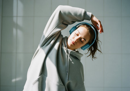 A serene young woman wearing a cozy gray hoodie and blue headphones performs a deep side stretch, enjoying music and movement in a sunlit indoor space. this image conveys wellness and relaxation.の素材