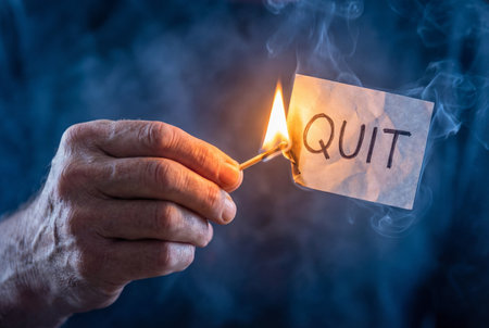 An older male hand holds a flaming wooden matchstick to set fire to a small white piece of paper with the text quit written in black ink against a smoky dark blue background.の素材