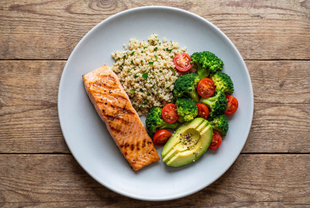 A healthy lunch plate featuring grilled salmon fillet served with fluffy quinoa salad, steamed broccoli florets, cherry tomatoes, and fresh avocado slices on a rustic wooden background.の素材