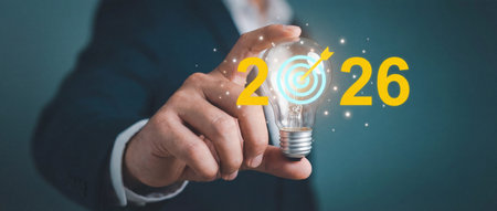 Businessman hand holds a glowing light bulb featuring the year 2026 with a target icon to symbolize strategic goals and corporate innovation planning for future success.の素材