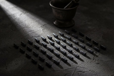 Meticulously arranged black medical capsules sit in a grid pattern on a dark textured concrete surface under a single ray of light with a vintage mortar and pestle in the shadow.の素材