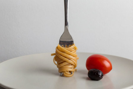 Silver fork stands upright holding a neat coil of cooked linguine pasta on a white plate accompanied by a single red cherry tomato and a black olive against a plain background.の素材