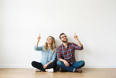 Young couple sitting on the floor leaning against a plain white wall and pointing upwards with smiles on their faces in a relaxed and happy mood in an empty roomの素材