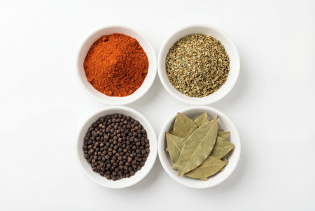 Top view of four white ceramic bowls arranged in a square containing red ground paprika dried oregano black peppercorns and green bay leaves on a plain white background for culinary use.の素材