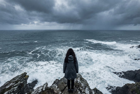 A solitary woman in a dark raincoat stands on a rugged cliff edge gazing at the turbulent ocean waves and white foam under a dramatic moody storm sky.の素材