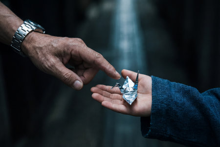 An elderly hand with a metal watch points gently toward a delicate silver origami crane resting safely in the small open palm of a young child wearing a textured blue sleeve.の素材