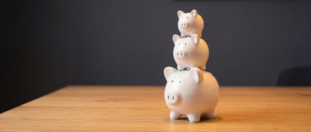 A vertical stack of three white ceramic piggy banks ranging from large to small stands on a wooden table against a dark gray background to represent increasing savings and financial investment growth.の素材