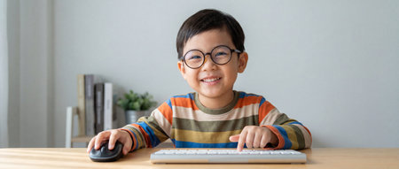 A cheerful little asian boy with round glasses and a striped shirt sits at a light wooden desk while typing on a white keyboard and holding a black mouse during an online lesson at home.の素材