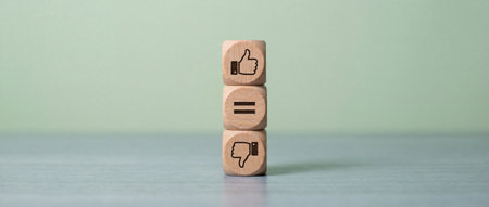Three wooden cubes are stacked vertically showing thumbs up equals and thumbs down symbols on a light background representing approval rating comparison and user feedback in a minimalist settingの素材
