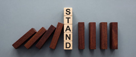 A vertical stack of light wooden cubes spelling the word STAND stops a row of falling dark wooden blocks from knocking over the remaining pieces in a domino effect concept on grey.の素材