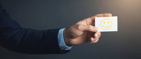 A businessman in a dark suit holds a small white paper card featuring a yellow smiley face drawing which represents customer satisfaction and positive service feedback on a grey background.の素材