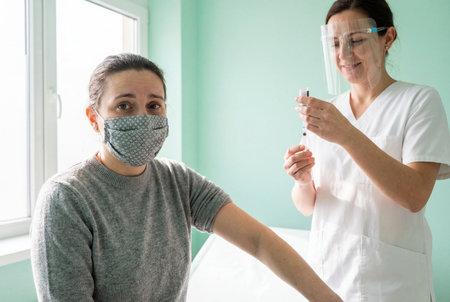 A female patient wearing a protective face mask sits with her arm exposed while a smiling nurse in a face shield prepares a syringe for vaccination in a bright green medical clinic room.の素材