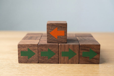 One unique wooden cube with an orange arrow facing the opposite direction stands on top of green arrows moving forward to represent nonconformity and individuality.の素材