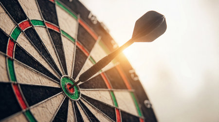 A sharp metal dart striking the bullseye of a standard dartboard in warm light symbolizing the successful completion of a targeted business goal or strategy.の素材
