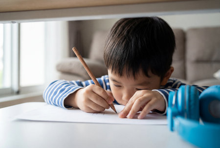 A concentrated young asian boy in a striped shirt sits at a white table holding a wooden pencil to draw on a sheet of paper while blue headphones rest on the surface in the foreground at home.の素材
