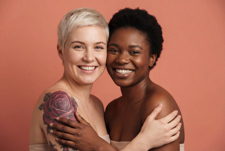 Two beautiful young women of different ethnicities standing cheek to cheek and hugging warmly with bare shoulders while smiling at the camera against a solid peach colored studio backgroundの素材