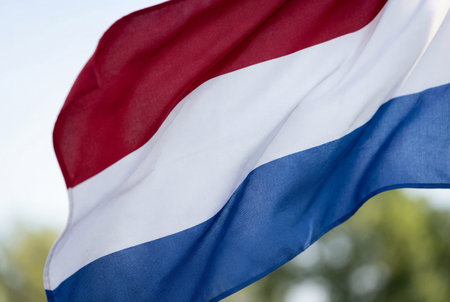 A close up view of the dutch national flag waving in the wind featuring horizontal stripes of red white and blue fabric against a blurred natural background of sky and green trees.の素材