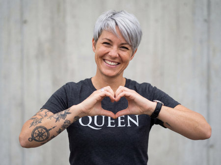 A happy middle aged woman with short silver hair and tattooed arms stands before a concrete wall while making a heart symbol with her hands and smiling brightly.の素材