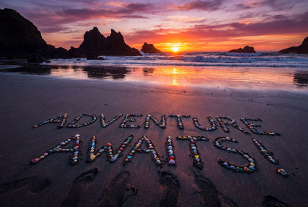 The words adventure awaits are spelled out on the wet sand using colorful seashells and pebbles against a backdrop of crashing waves and jagged sea stacks under a vibrant orange and purple sunset.の素材