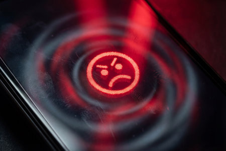 Macro view of a glowing red unhappy face icon on a smudged smartphone screen with a ripple effect symbolizing cyberbullying, negative feedback, digital error, or online dissatisfaction.の素材