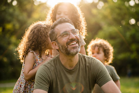 A happy bearded father wearing glasses smiles broadly while three curly haired children play and whisper behind him in a sunny green park during summer.の素材