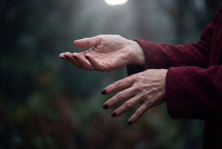 Close up view of wrinkled hands of a mature woman with dark red nail polish reaching out to catch falling rain drops against a blurred dark green forest background during a rainy day.の素材