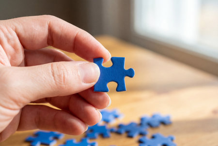 Hand holding a single blue jigsaw puzzle piece over a wooden table with scattered pieces, representing the concept of finding a solution and completing the game with logic and strategy.の素材