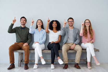 Five happy young multiethnic friends in smart casual clothes sit on a wooden bench against a grey wall while pointing their index fingers upward at empty copy space above their heads.の素材